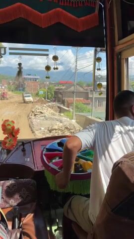 The video is filmed from inside a bus, looking out through the windshield. A person's hands are visible operating the steering wheel, which is decorated with colorful fabric fringe. The bus is on a muddy, unpaved road that appears to be under construction or repair. Buildings and trees line the road, and mountains are visible in the background under a cloudy sky. There are decorative items inside the bus, including artificial flowers and Buddhist iconography. Several other vehicles, including a white van and a red vehicle that may be a tractor, are visible on the road ahead. The overall scene suggests travel through a rural or developing area with challenging road conditions.