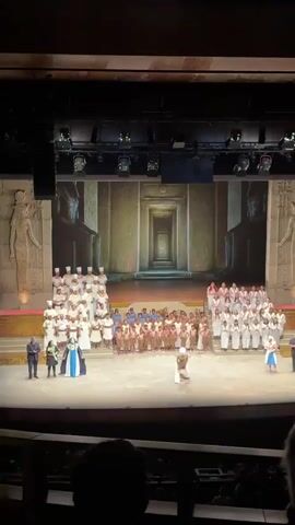 A wide-angle shot captures the finale of what appears to be a stage production. A large cast is assembled on a stage decorated with what looks like Egyptian motifs, including projected images of statues and doorways. Most of the cast is dressed in white robes or simple tunics. Four individuals stand at the very front of the stage, two on each side, dressed in more elaborate costumes. The audience applauds enthusiastically as the performers take their bows. Overhead lighting illuminates the scene, and the video is shot from the perspective of someone seated in the audience.
