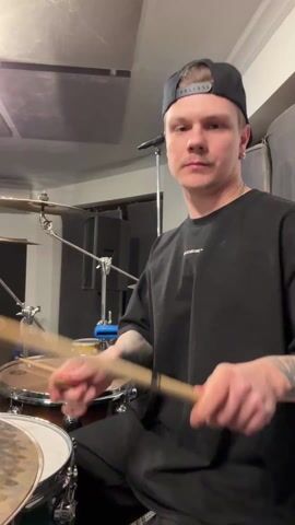 A man is seen playing a drum set in what appears to be a recording studio. He is wearing a black t-shirt and a black baseball cap. He has short, light-colored hair and visible tattoos on his left arm. He is holding drumsticks and striking the drums, cymbals, and other percussion instruments with noticeable energy and facial expressions. The drums are brown and white, and the cymbals are silver. In the background, there is soundproofing material on the walls, as well as a speaker and some other musical equipment. The lighting is bright.