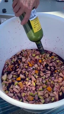 A person's hands are seen pouring red wine from a dark green bottle into a large white tub filled with chopped apples and oranges. The tub is brimming with the fruit and the dark red liquid, suggesting the preparation of a large batch of sangria or punch. The wine bottle is partially obscured by the person's hands but its label is visible, featuring a yellow sticker with Spanish text. The background shows a table with several other bottles, some with caps, and a glimpse of a bright, outdoor setting, potentially a picnic or outdoor gathering, indicated by the sunlight and checkered tablecloth. The scene appears to be taking place during the day, likely outdoors given the lighting. The overall impression is one of casual, communal food or drink preparation.