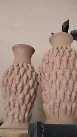The camera pans out, showing two unglazed, undecorated clay pots of similar shape and design resting on a shelf. The pots are beige and decorated with what appears to be numerous small, similarly-shaped clay shapes. To the side of the pots are several other ceramic art objects, including a yellow vase and what appears to be a rock. The pots are placed against a white wall and the shelf is numbered.