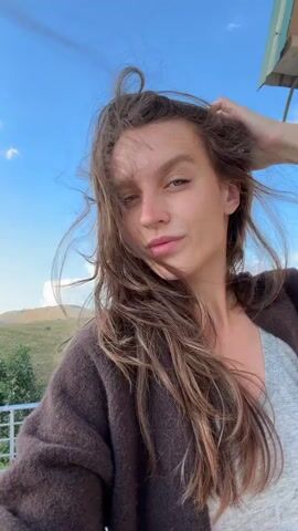 This media file is a selfie video of a young woman in an outdoor, mountainous setting. She has long, brown, wavy hair that is being blown by the wind. She is wearing a dark brown cardigan over a light grey top.

The scene appears to be a scenic overlook or a viewpoint, with rolling hills and a winding road visible in the background. The sky is blue with scattered white clouds. The time of day seems to be late afternoon or early evening, as indicated by the soft lighting.

The woman is looking at the camera and adjusting her hair, smiling and looking relaxed. She appears to be enjoying the natural beauty of her surroundings. There is no visible text in the video.