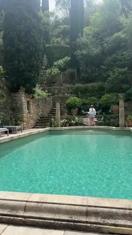 The video shows a man diving into a turquoise swimming pool on a sunny day. At the beginning, the man, wearing dark green shorts, is seen leaning over the pool edge, poised to dive. In the background, a woman in a white top and light trousers stands near the far side of the pool. The setting is an idyllic outdoor area with lush greenery, tall cypress-like trees, and ancient-looking stone architecture, including terraced walls and steps. Potted plants and outdoor furniture are visible around the pool area.

The man then dives into the pool, creating a large splash. Another woman, dressed in a white dress, is sitting on a bench to the left of the pool, observing the scene. After the dive, the man is visible swimming just below the water's surface. The bright natural light indicates it is daytime with clear weather. The surroundings suggest a private estate or villa, possibly in the French countryside given the provided location context of Saint-Victor-la-Coste, France, with its characteristic stone work and abundant vegetation. No text is visible in the video.