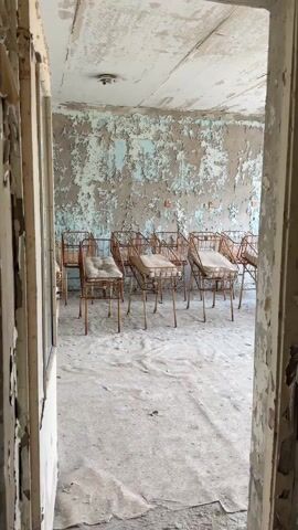 A woman with long brown hair walks into a room through a dilapidated doorway. The paint on the walls of the room is peeling and faded. There are several chairs with wire frames and cushions lined up against the back wall of the room. The floor is covered in debris. The door frame is badly damaged, with layers of peeling paint. The overall impression is one of neglect and decay.
