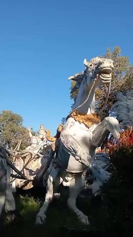 The video pans across a statue of a white horse with ornate golden decorations. The horse is in a rearing position, with its mouth open. Behind it, another statue of a horse and chariot can be seen, with a figure seated in the chariot. The statues are located outdoors, with a clear blue sky and some trees and greenery visible in the background. The overall impression is of a monument or a garden sculpture.