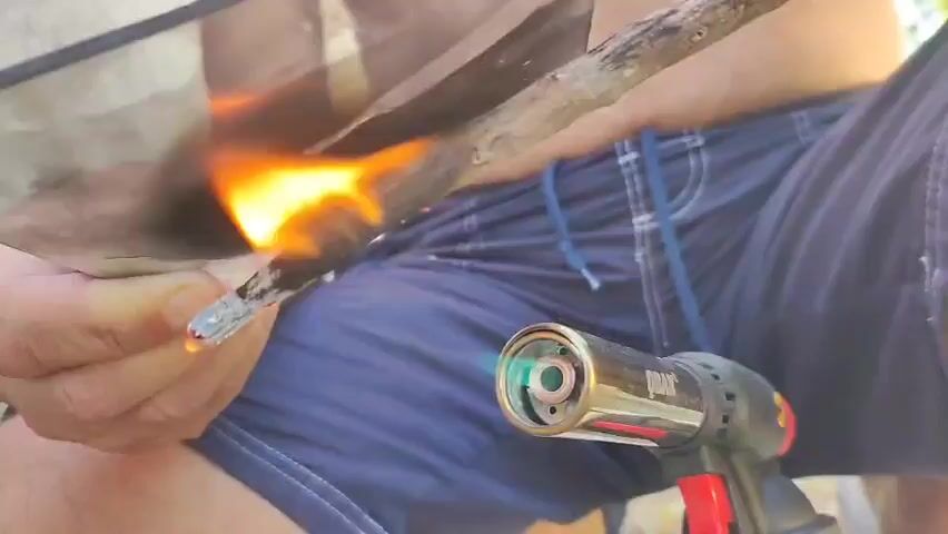 A close-up shot shows a person holding a small stick over an open flame coming from a butane torch. The stick is burning and dripping some liquid. A reflective surface is held to the left of the stick, catching the flame. The person's hands and legs wearing blue shorts are visible. The background is blurry and appears to be outdoors.