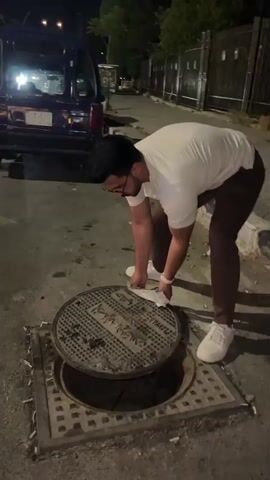 A man is shown in an outdoor evening setting. He is leaning over a manhole cover, cleaning it with a paper towel. The man is wearing glasses, a white t-shirt, brown pants, and white shoes. The manhole cover is round and sits within a square frame on a paved street. A blue car is parked nearby. There are buildings and a fence in the background.