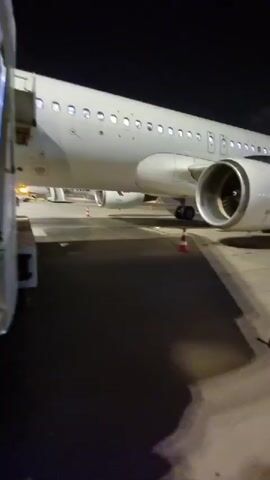 The video shows a group of people walking up a portable walkway to board an airplane at night. The walkway is covered and has a curved roof. The people are dressed in casual clothing and are carrying bags. The airplane is white and has several windows. The walkway is attached to the plane, allowing passengers to enter. The people ahead on the staircase appear to be entering the aircraft.
