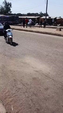 The video depicts a street scene in what appears to be a developing area, possibly a town or city in Africa. Initially, a person on a white motorcycle pulls onto the road from the side, adjacent to a dark-colored sedan. The camera then pans to the right, showing a man pushing a wheelbarrow heavily loaded with a large sack down the road. Two individuals, one carrying a red suitcase and the other with a long object (possibly a broom or a similar tool), walk along the sidewalk. There are other people visible in the background, along with parked vehicles, buses, and what appear to be market stalls. The road surface is worn, and the overall environment suggests a bustling, populated area with active transportation and commerce. The sky is clear and the weather appears to be sunny.
