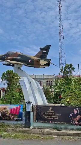 The video shows an outdoor scene with a monument of a camouflaged jet airplane mounted on a pedestal. The airplane is positioned against a sky with scattered clouds. In the background, there are several buildings and a tall communication tower. A large billboard with advertisements is visible in the foreground, featuring a man's face and text in Indonesian. The surrounding area appears to be grassy with some debris scattered on the ground. The camera pans slightly to the right, showing more of the environment.
