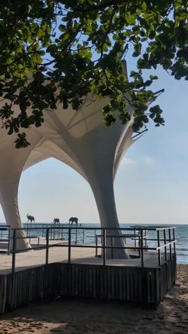 A fixed-perspective daytime shot shows a pavilion-like structure with a white tent roof, four arched pillars, and a dark-colored wood railing. The structure sits on a concrete platform that stretches out to the sea, visible in the background. On the platform, there are three metal elephant statues on stilts facing the water. The sky is blue with some light clouds. In the foreground, the leaves of a tree partially obscure the view. Music plays in the background.