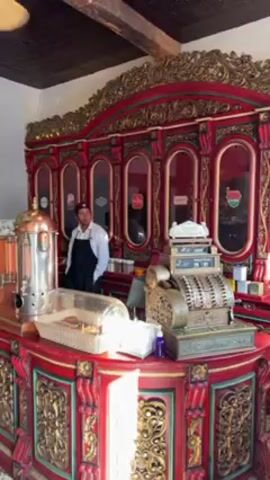 The video shows the interior of what appears to be a coffee shop or cafe with a vintage aesthetic. A person in a white shirt and black apron stands behind a highly ornate, red and gold counter. On the counter are a copper coffee urn, a display of pastries, and an antique cash register. The background features a red and gold display case with glass windows, possibly containing coffee or tea. The camera pans to the right, showing a tiled floor, another section of the counter with a coffee machine and cups, and a doorway with a person standing in it. The camera continues to pan, revealing a "Souvenir" shelf stocked with jars, followed by a vintage-looking piece of machinery stirring something in a copper vessel. The overall ambiance is old-fashioned and decorative.