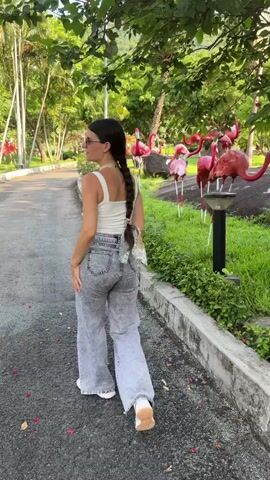 A young woman with dark hair in a braid, wearing a white tank top, acid-wash jeans, and sunglasses, stands on a paved path in a park-like setting with palm trees and lush greenery. She is facing the camera and poses with her hand on her hip. In the background, there are numerous pink flamingo statues of various sizes. The woman turns and walks away from the camera, then turns back and stretches her arms upwards. She then turns and walks away again.
