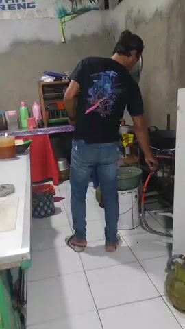 A man stands in a kitchen, facing away from the camera. He wears a black t-shirt with a graphic design on the back and blue jeans. He is standing next to a stove with a pan on it. He appears to be cooking or preparing to cook. The kitchen is sparsely furnished with concrete walls and tiled floors. There are various items visible on shelves and counters, including bottles, containers, and cooking utensils. The lighting is dim, suggesting it may be evening or the kitchen is not well-lit.