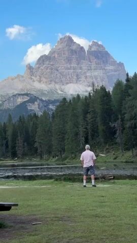A man stands facing away from the camera, looking at a large mountain peak. He is wearing a pink shirt and grey shorts. The man is in a grassy field next to a body of water. A line of trees is behind the water, and the mountain peak is visible above the trees. The sky is blue with a few white clouds. Another person is visible to the right of the frame, also facing away from the camera.