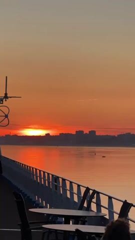 The video shows a sunset over a body of water, with a city skyline in the distance. The sun is low on the horizon, casting a warm orange glow across the sky and water. The foreground shows the deck of a boat, with tables and chairs set up. A railing runs along the edge of the deck, and some equipment is visible on the left side of the frame. There are no people clearly visible in the video, although there might be a person or two in the foreground, mostly out of focus. The scene appears peaceful and serene.