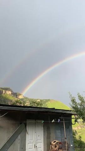 The video shows a double rainbow against a grey sky. Below the rainbow is a green-covered rooftop with some sort of building structure on it. There are two white doors visible on the front of the building. On a wire strung across the roof, there are three clothes pins with two of them clipped to the wire. To the right of the roof is a tree. In the background, there are green hills.