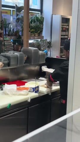 The video shows a person wearing a black chef's uniform and a black hat preparing food behind a counter in what appears to be a store or restaurant kitchen. The person is wearing white gloves and is in the process of wrapping food in white paper. On the counter in front of them, there is a package of cream cheese, a container of butter, a red cutting board, and what looks like sliced salmon. To the left of the frame, another person wearing a green shirt and a dark baseball cap is visible, facing away from the camera and towards shelves stocked with plants and other items. The environment appears to be a brightly lit commercial space.