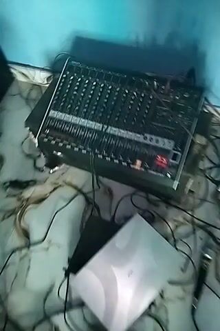 The video displays an audio mixing board with multiple sliders and knobs. Cables connect to the mixing board, leading to other equipment. A large speaker with three distinct circular components is visible, also connected by cables. The scene appears to be indoors, with a light blue wall in the background and a patterned floor. The overall impression is of a setup for sound amplification or recording.