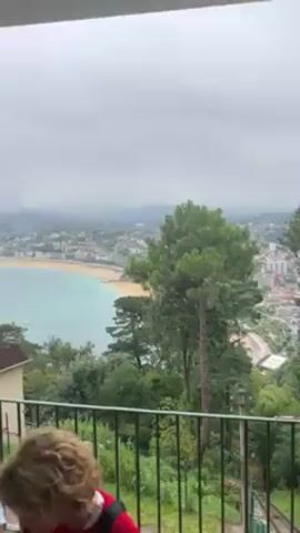 The video displays a scenic view from a balcony. In the foreground is a black metal railing, with lush green vegetation beneath it. The main focal point is the panoramic vista of a city nestled along a coastline. The beach is a light sand color, and the water is a pale turquoise. In the background, buildings and structures can be discerned, partially obscured by a hazy sky. The overall atmosphere is tranquil and scenic, with a touch of serenity.