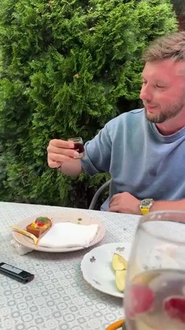 A man with short brown hair and a beard, wearing a light blue t-shirt and a yellow watch, sits at a table outdoors. He holds a small shot glass filled with a dark liquid to his lips and drinks it. In front of him on the table is a plate with a piece of food topped with what appears to be caviar and a white napkin, next to a cell phone. To the right of that plate is another plate with sliced fruit. A glass with red berries is partially visible in the foreground. The man is sitting in front of a green hedge.