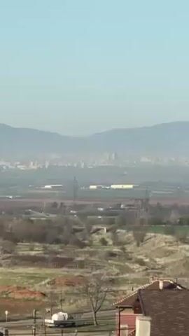 The video pans up across a landscape of fields and trees, revealing a city in the distance with mountains behind it.  The tops of the mountains are capped with snow.  There are power lines in the fields below, and a road is visible near the bottom of the frame.