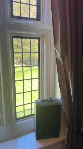 A camera pans across a series of six-paned, black framed windows. The windows look out onto a large green lawn with mown lines and mature trees. The window frames are white with gothic revival details. A brown suitcase and a small, antique clock sit on the windowsill.  A brown curtain hangs to the right of the windows. There is brick visible through a window in the distance.