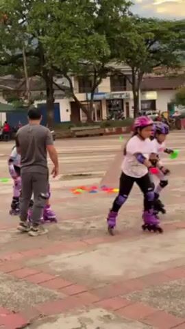 A small girl wearing a helmet and knee pads skates around a brick-paved plaza, weaving between orange and green cones. She has colorful decorations on her arms and legs. A man, presumably her coach, is next to her giving instructions. Another child is visible to the right also skating. A massive tree with a large canopy dominates the background. In the far background are buildings and a mountain range under a cloudy sky. Several other people are milling around the plaza, some are seated on benches. A sign that says "YO AMO TIMAYA" is partially visible. The skater is working on her technique as she navigates the cones.