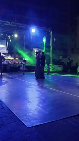 A man and woman are dancing in the center of an outdoor stage area at night. The man is wearing a dark shirt and jeans. The woman is wearing a dark, floor-length dress. They are holding each other and appear to be performing a choreographed dance. The stage is illuminated with bright, colored lights, primarily green and purple, which cast shadows on the ground. In the background, there is a stage with equipment and lighting rigs, as well as some people standing and watching. There are also barriers and some buildings visible in the distance. The ground is paved with cobblestones and covered with a large, rectangular reflective surface, likely a temporary stage or dance floor. The overall atmosphere suggests an outdoor event or performance.