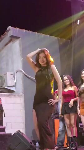 A young woman in a long black dress with a high slit is dancing on a stage. She has her hand on her hip and her other hand is raised above her head. She is smiling and looking at the camera. There are other people on the stage behind her, including two other women in dresses and a man in a denim jacket. There are also speakers and stage lights visible. The background appears to be a building with a corrugated roof. The lighting is dim and colorful.