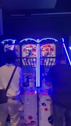 Two people are playing basketball arcade games in a dimly lit space with blue lights. On the left is a person with curly hair wearing a white long-sleeved shirt and white pants with a brown bag slung across their shoulder. On the right, a person in a black shirt and dark pants is also playing the game. Both individuals are actively throwing basketballs into the hoops of their respective machines. The arcade games are brightly colored with depictions of Sonic All-Stars.