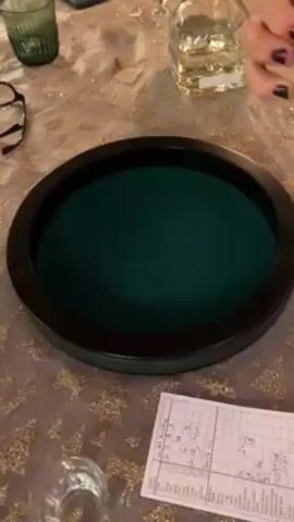 A hand shakes five dice in a light brown cup, then drops them into a round, green-felt-lined dice tray with a dark brown rim. There is a scorecard with handwritten numbers and text lying next to the tray. Beside it is an empty glass, a glass with liquid in it, and a bottle of clear liquid. Eyeglasses are on the table to the left. The table has a gold-colored patterned tablecloth.