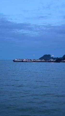 The video shows a wide shot of a body of water, likely the sea, under a twilight sky. In the background, there are several large boats or ferries docked near a shoreline with lush green hills and mountains. The sky is a deep blue with scattered clouds, and there are a few lights visible on the shore and on the boats. The water is calm, with gentle ripples reflecting the dim light. The camera appears to be moving slowly, offering a panoramic view of the scene. There are no people visible in the frame. The overall impression is one of a peaceful evening at a harbor.