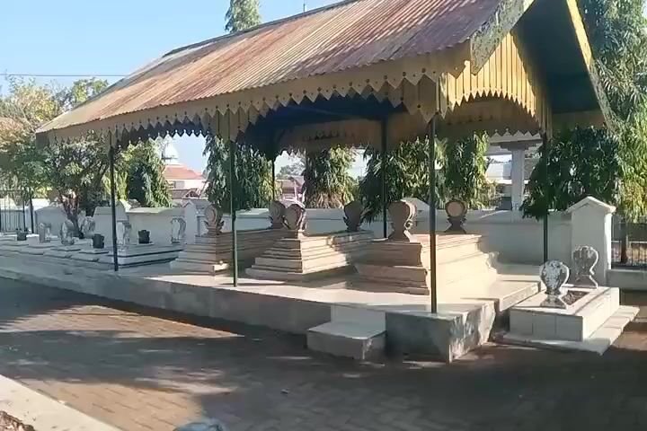 A graveyard is shown with several rows of graves under shelters. The graves are made of stone and many are rectangular. Some graves have ornate tops. The shelters have metal roofs and decorated trim. The ground is paved with bricks and there is dirt around the graves. Trees are visible behind the graves and in the background. The sky is blue and the sun is shining, casting shadows. There are no people visible in the video.