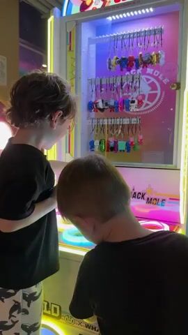 Two boys are playing a whack-a-mole style arcade game. The game machine is brightly lit with neon lights in pink, yellow, and blue. Above the game area, a display case holds an assortment of small, colorful toys on hooks, likely prizes. The game itself features multiple illuminated targets that light up as players attempt to hit them with a soft mallet.

The younger boy, in a black t-shirt, is reaching towards the control panel of the game, possibly to insert coins or select options. The older boy, wearing a black t-shirt and patterned shorts, is holding the mallet with both hands, poised to strike a target. His focus is on the game screen, as is the younger boy's.

The scene appears to be indoors, likely an amusement arcade or entertainment center, given the nature of the game and the surrounding bright lighting. The backdrop suggests a playful and energetic environment. There is no indication of time of day or weather. The emotional atmosphere seems to be one of focused engagement and fun.

Visible text on the machine includes "WHACK-A-MOLE" in large letters, along with Cyrillic text that appears to be instructions or game information. There's also text indicating a price, "200 p," likely referring to the cost to play. The overall impression is a snapshot of childhood enjoyment at an arcade.