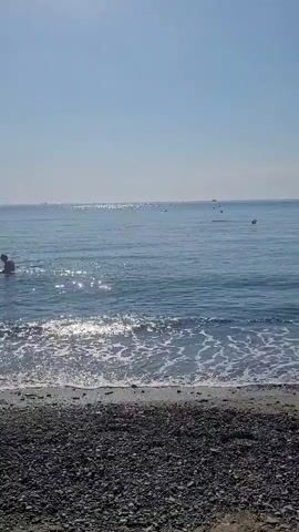 The video shows a view of the ocean from a pebble beach. The water is blue and calm, with small waves lapping at the shore. The sun is shining, and there are some small boats or buoys visible in the distance. The sky is clear and blue. The foreground of the video is a beach covered in dark grey pebbles.