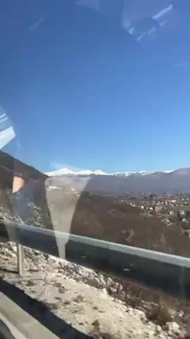 The video starts with a view from a moving vehicle of a landscape with mountains in the distance, some snow-capped. The vehicle then passes by a rocky terrain with sparse trees. The video then shifts to an interior view, with a blurry white object visible. The camera then points down, revealing the same rocky terrain. The white object is now clearly a garment or fabric lying among the rocks.