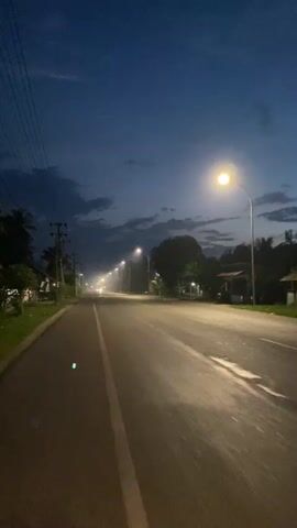 The video captures a dimly lit asphalt road at dusk or dawn. Streetlights spaced out along the right side of the road illuminate the scene, creating a hazy glow that stretches into the distance. The sky is a deep blue with scattered clouds. Power lines are visible on the left side of the road, running parallel to it. Trees and some small buildings can be seen on either side of the road, partially obscured by the low light and haze. There are no people or vehicles clearly visible in the frame. The road appears to be empty, with a white line dividing the lanes.