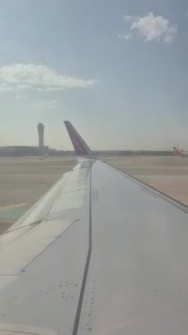 The video shows a view from an airplane window of the wing of the plane, with an airport control tower and other airplanes visible in the background. The sky is hazy with a few clouds. The WizzAir logo is visible on the tail of the plane. The plane appears to be on the ground. Sounds of people speaking are in the background.