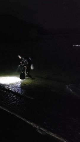 The video depicts a very dark scene with a person in scuba gear walking through shallow water with a bright light. Further in the distance, another person or object is visible, also emitting light. The environment seems to be a shoreline at night. The overall visibility is poor, limited by the darkness and the light sources.
