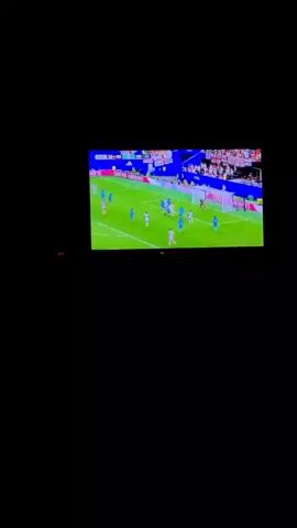 This media file shows a television screen displaying a soccer match. The game is taking place on a green field with spectators in the background. Advertising boards around the field display "QATAR AIRWAYS" and "LENOVO". The score board at the top left indicates the game is at 0-0 and the time is 00:00. The players are wearing white and blue uniforms. The scene appears to be indoors given the darkness of the surrounding area. The television screen is bright and clear, showing the action of the game in progress. The image zooms in on two players in white uniforms, one of whom appears to be celebrating a goal, while the other congratulates him. The mood seems celebratory and excited.