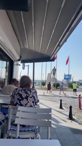 A static, eye-level shot taken from under an awning. The awning is a light grey retractable canvas structure. The foreground shows the back of a white table with blue and white striped placemats and chairs. To the left, a person is partially visible, wearing a blue and white floral top. In the middle ground, a paved walkway is visible, with a stone border and black bollards. Several people are walking across the walkway. Behind the walkway, a street with vehicles and trees is visible. On the far side of the street, a statue of a person is visible, standing on a pedestal. Two Turkish flags are flying on poles behind the statue. The sky is clear and blue. The lighting suggests it is daytime.