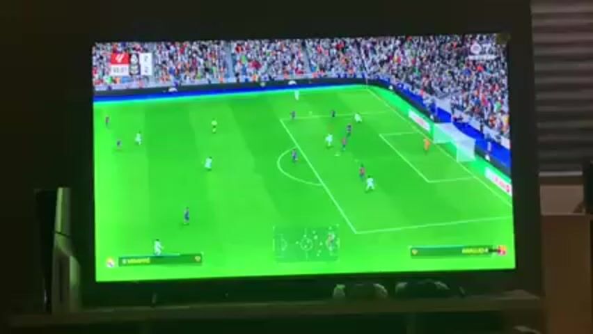 A TV displays a soccer video game, where a player tries to score a goal. The game shows players on the field, with spectators in the background. The perspective switches to a closeup of the goalkeeper arguing with another player. A PS5 is visible to the left of the TV, and two game controllers sit below the TV.