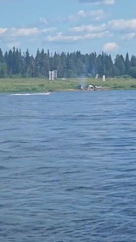 A jet ski is speeding across a wide body of blue water under a cloudy sky. On the far bank of the water, a dense line of green trees forms the horizon. In front of the trees, on a grassy shore, are several small structures, possibly tents or temporary shelters, and a few vehicles. There are also two white posts visible on the shore.