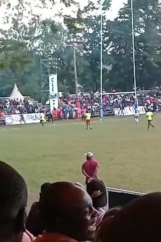 The video captures a rugby match unfolding on a grassy field, with a crowd of spectators watching from the sidelines and stands. Several players are visible in red and green jerseys, engaged in the game. In the foreground, the backs of people’s heads are visible, suggesting the perspective of someone in the audience. Behind the players, banners with "CLEO MALALAH" are displayed. In the background, a metal structure with "CLE" on it is partially visible among the trees. The video seems to be recorded from a mobile device.