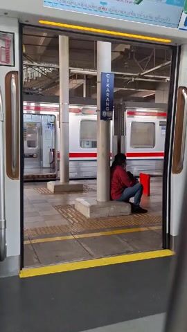 The video shows the interior of a train at a station. The doors of the train are open, revealing a platform with trains on adjacent tracks. A person is sitting on a bench on the platform, facing away from the camera. Another person, wearing a hijab and glasses, walks into the train. The video captures the moment the doors close.