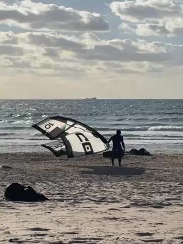 A person stands on a sandy beach holding a kite, with the ocean in the background. The kite is white and black with some green and writing, and it lies partially on the sand. The person is silhouetted against the bright sky, and there are bags lying on the sand near them. The ocean has waves, and a large boat is visible far away. The sky is partially cloudy.