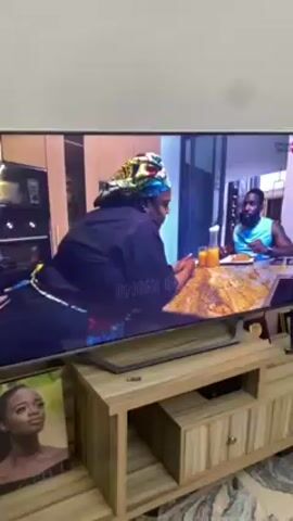 The video shows a flat-screen television displaying a scene of two people sitting at a table. The woman is wearing a colorful head wrap and a dark top. The man, wearing a light blue tank top, is eating with a fork. A pitcher of orange juice and glasses are visible on the table along with plates of food. The table has a brown and tan marbled surface. The television is sitting on a light brown wooden stand. A framed portrait is to the left of the TV stand. The environment appears to be inside a home, with a neutral colored wall in the background.