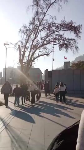 The video begins with a view of a sidewalk with people walking on it, with a large tree casting shadows. In the background, there is a wall and a building with a flag. The camera then pans to the right, showing a statue of an angel with wings. More people are seen in the background, some sitting and others walking. The scene then transitions to a wide shot of a park-like area with trees, streetlights, and people walking. Finally, the camera focuses on a tall, modern skyscraper under construction, with cranes visible. People are seen relaxing on the grass in the foreground, and two individuals are walking together. The sky is clear and blue throughout the video.