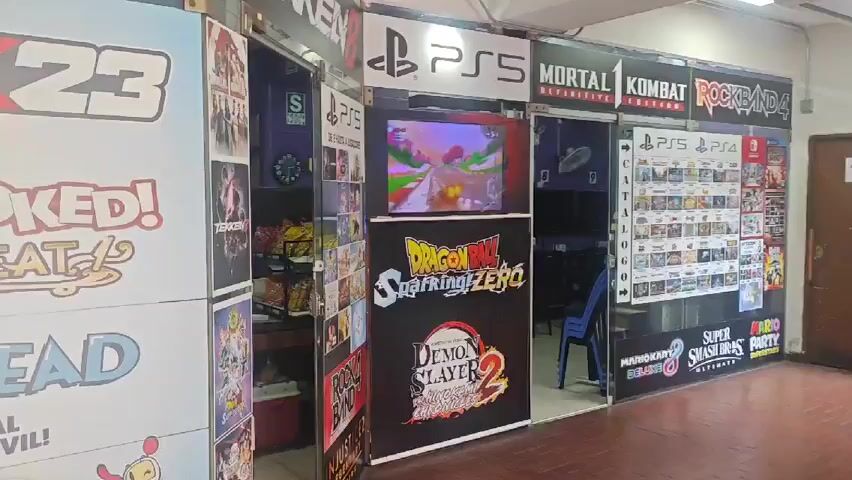 The video shows storefronts in what appears to be an indoor shopping center. There are video game and anime themes present. Some storefronts display posters of popular games like WWE 2K23, Cuphead, and Dragon Ball Sparkling Zero. Some stores are decorated with figures of anime characters like Luffy from One Piece and Ash from Pokemon. People can be seen browsing in and around the stores. One person is seated inside a shop, possibly reading or working. The setting is well-lit and appears to be a commercial area focused on entertainment and related merchandise.